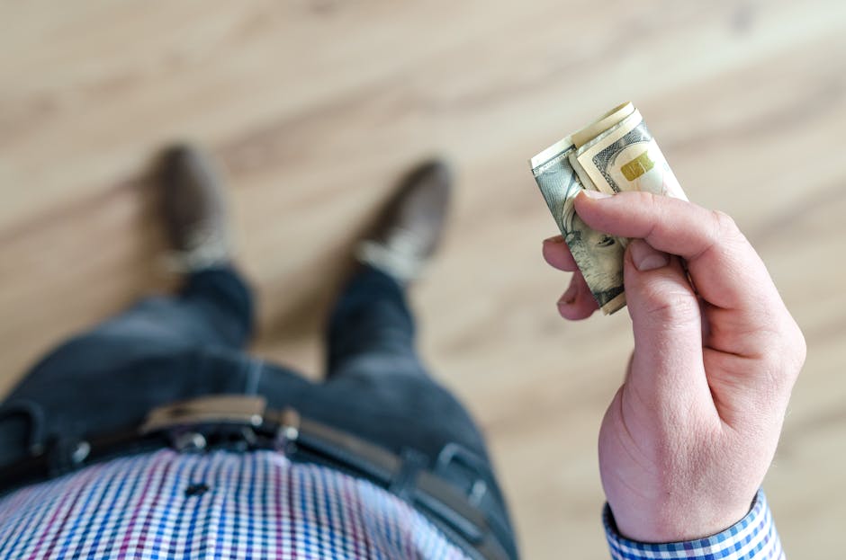 A person wearing denim and a checked shirt holds folded dollar bills in their ha