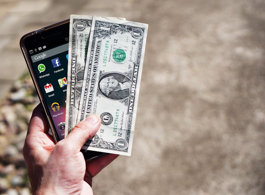 Close-up of a hand holding US dollar bills and a smartphone outdoors, showcasing