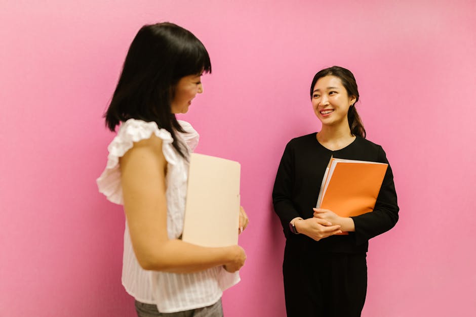 Two Asian women in business attire discussing work while holding folders against