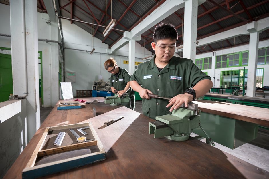 Two workers in a well-lit Indonesian workshop using clamping tools and safety ge