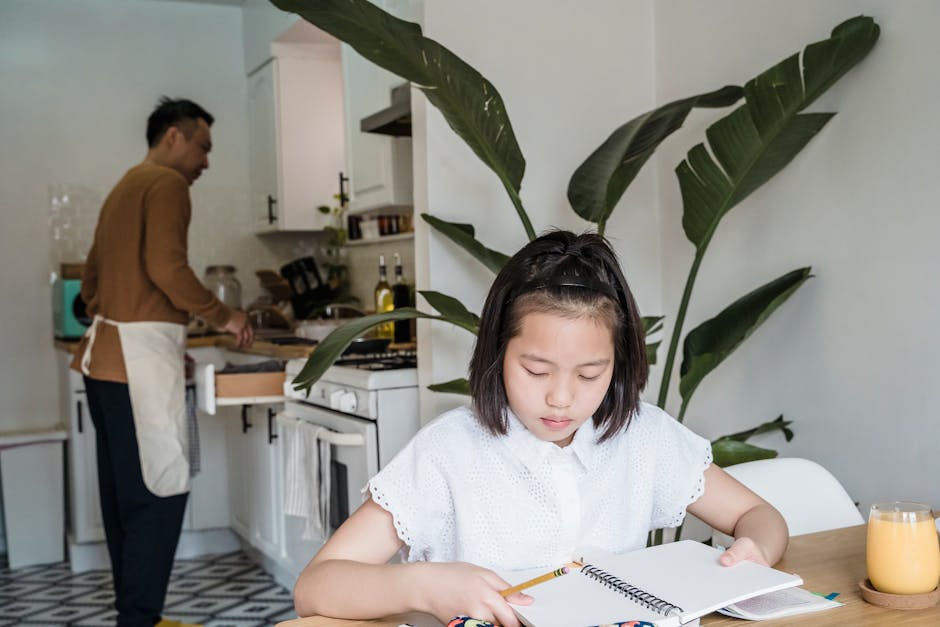 A young girl focuses on her homework while her parent cooks in a cozy kitchen se