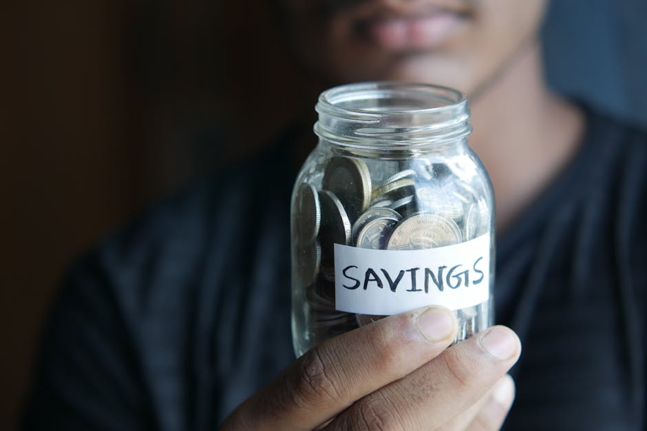 A close-up image of a person's hand holding a jar full of coins labeled 'Savings