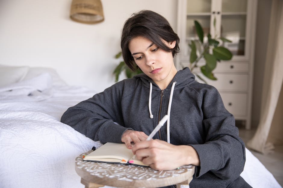 Young woman in a hoodie writing in a notebook at home, capturing a reflective mo